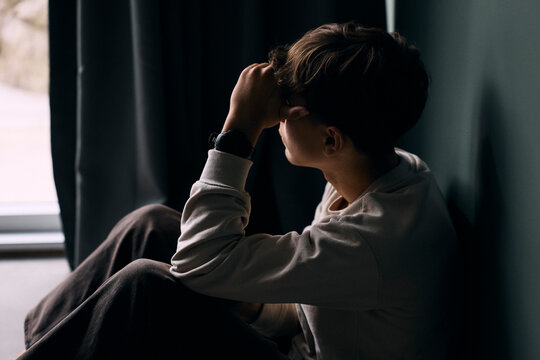 Lonely teenage boy with head in hand sitting by wall at home