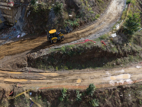 Aerial view of a yellow backhoe loader working on a muddy dirt road at a construction site with excavated earth and sparse vegetation in Istanbul, Istanbul, Turkiye.