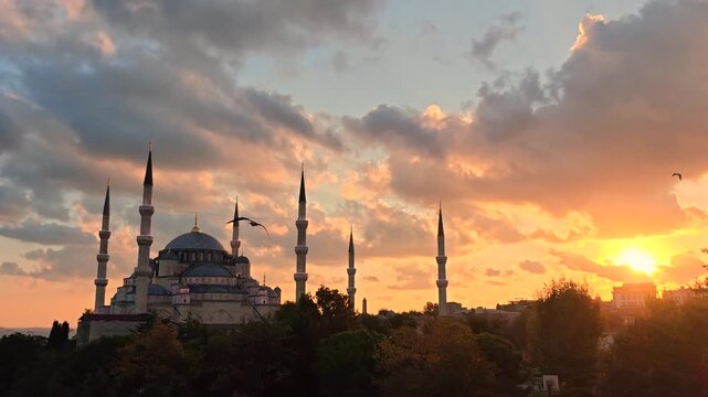 Backlit silhouette of mosque with central dome and six tall minarets against glowing sunset clouds, with birds flying across sky and buildings fading into shadow.