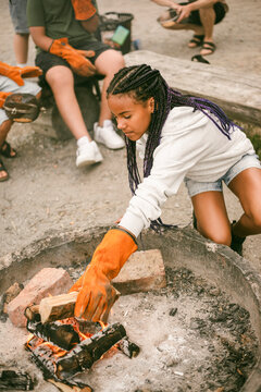 Girl with braided hair wearing gloves while putting firewood into small campfire