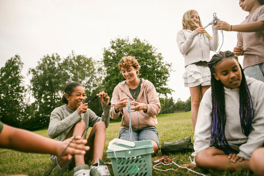 Happy girl learning rope tying skills with female leader while sitting on grass at summer camp