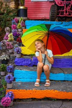 Young girl with rainbow umbrella sits on colorful steps surrounded by vibrant flowers in a lively outdoor setting