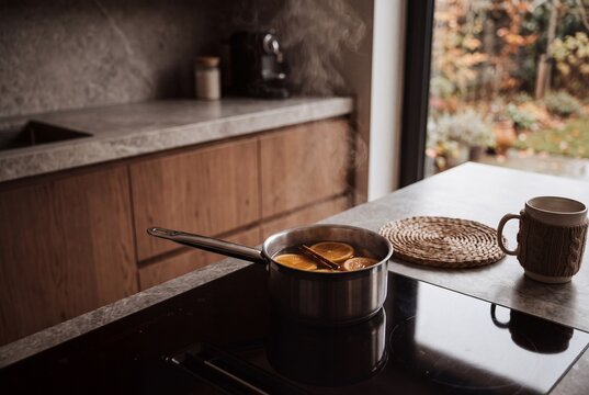 Steaming saucepan with orange slices and cinnamon on a modern kitchen stovetop. Hot autumn beverage preparation. Mug with knitted cozy on the counter