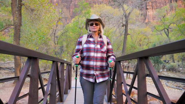 Active woman 50 59 years with trekking poles and backpack walking towards the camera on metal bridge in forest. Female hiker enjoying healthy outdoor lifestyle and adventure in national park zion
