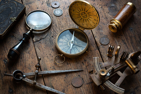 Antique navigation and exploration still life centered around an old compass on a rustic wooden surface, surrounded by an astrolabe, dividers, antique book, coins, spyglass, and magnifying glass. 