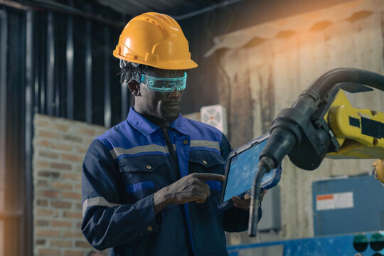 A professional Black male technician wearing glasses high-tech gear with a blue glow is touching a tablet screen; the background is a blurred view of a machinery room.