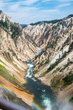 Aerial view of the Grand Canyon of the Yellowstone with the Yellowstone River flowing through deep rocky cliffs, pine trees, and a faint rainbow in the mist in Cody, Wyoming, United States.