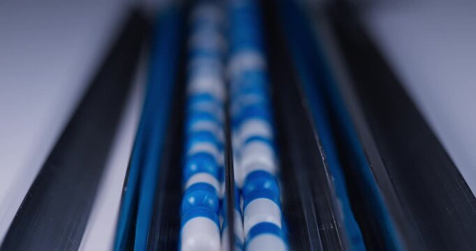 Tracking shot of blue and white capsules moving along stainless steel conveyor in pharmaceutical production line.