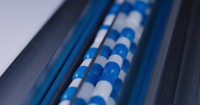Tracking shot of blue and white capsules moving along stainless steel conveyor in pharmaceutical production line.