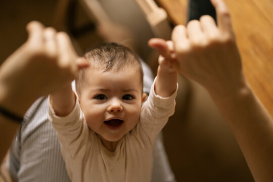 Baby smiles brightly while holding hands with adults in warm home setting