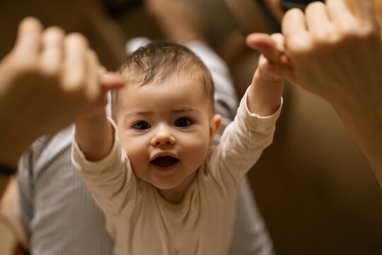 Baby smiles with arms raised as hands reach toward them in warm indoor setting