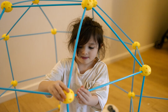 Young girl builds colorful geometric structure with focused expression indoors on wooden floor