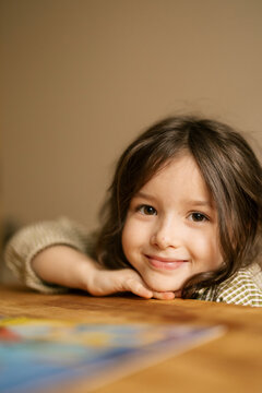 Young girl smiles while resting her chin on a wooden table in warm cozy room
