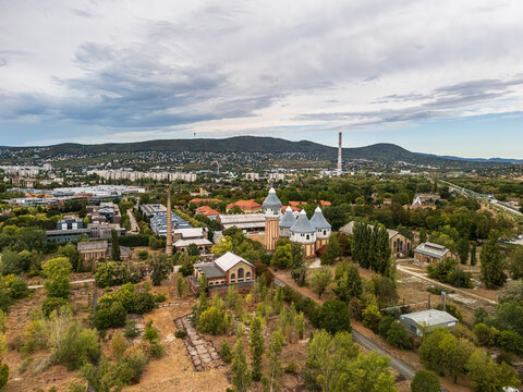 Aerial view of the historic Obuda Gasworks, with brick industrial buildings, towers and chimneys amid trees, urban development and the Buda Hills in Budapest, Hungary.