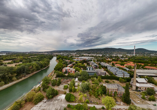 Aerial view of offices and industrial buildings beside the Danube and tree-lined Obudai Island in Budapest, Hungary, with Buda Hills beneath a cloudy summer sky.