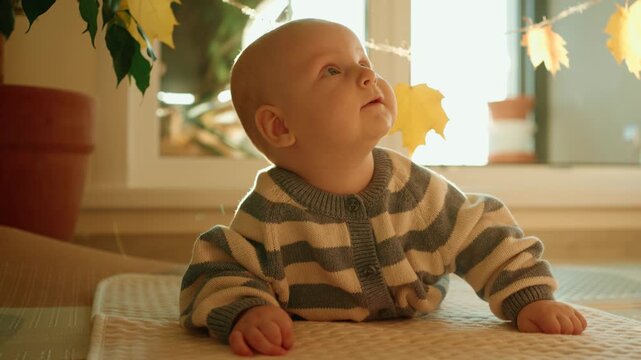 Cute infant in a striped sweater doing tummy time on a soft mat, playing with a garland of autumn leaves held by his mother's hand in a sunlit room, fostering early development