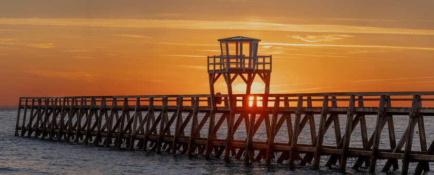 Luc-Sur-Mer, France - 08 08 2026:  Panoramic view of a wooden pier in front of the beach, sea, a colorful sunrise and Le Havre city far away