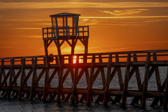 Luc-Sur-Mer, France - 08 08 2026:  Panoramic view of a wooden pier in front of the beach, sea, a colorful sunrise and Le Havre city far away