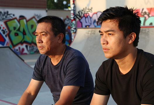 Generational gap and family bonding, asian father and son sitting together at a skatepark with urban graffiti background during golden hour sunset