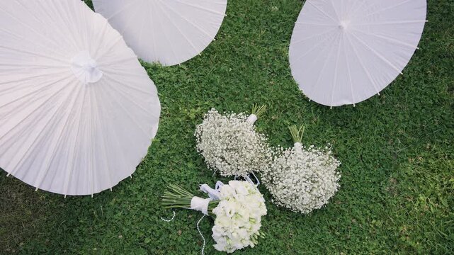 Wedding bridal bouquets and white paper parasols on the green grass, elegant wedding flower arrangements with gypsophila and white sun umbrellas outdoors, romantic marriage ceremony details.