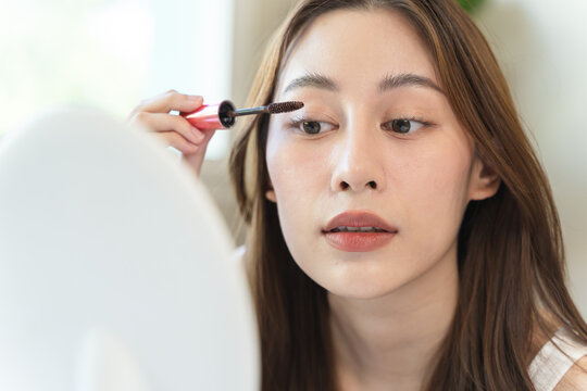 Beautiful young Asian woman applying brown mascara to her eyelashes in front of a mirror, daily makeup routine and morning beauty concept at cozy home.
