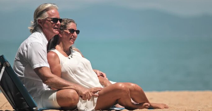 Senior couple in white enjoying a tropical Samui beach vacation, sitting on sand as the woman points toward the calm turquoise ocean and sunny blue sky, smiling and relaxed. Travel, holidays, relax
