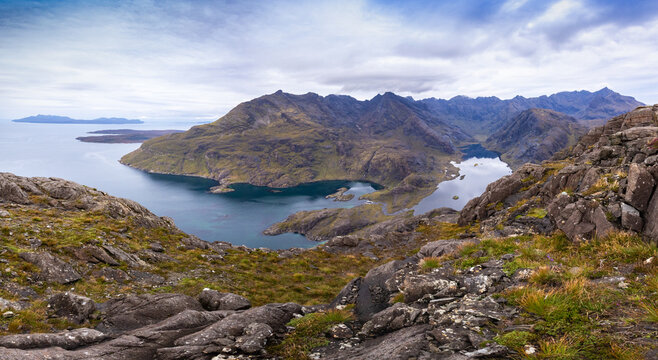 View of Sgurr na Stri overlooking the dark waters of Loch Coruisk with rugged rocky terrain and distant mountains under a cloudy sky in Isle of Skye, Scotland, United Kingdom.