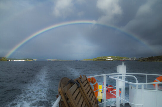 View of a vibrant rainbow arching over the sea with the Skye Bridge in the distance, seen from the deck of a boat under a dramatic stormy sky in Isle of Skye, United Kingdom.
