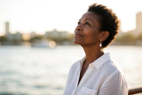 Thoughtful middle aged African American woman looks up towards sky. Mature black lady wears white linen shirt, pearl earrings. Stands near sea with city skyline background during golden hour.