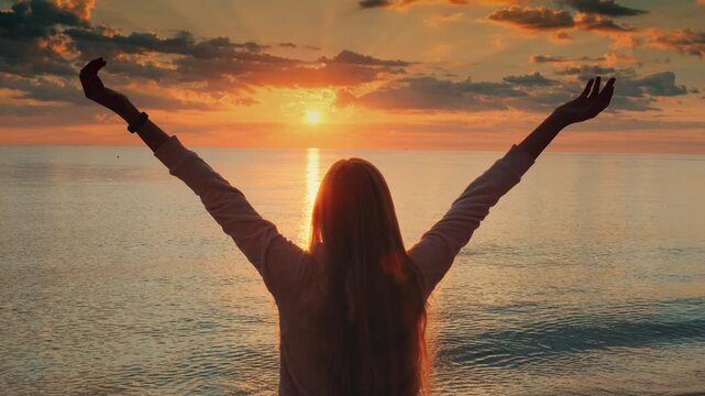 A young woman with long dark hair stands with outstretched arms facing the ocean at the beach during daytime, surrounded by sea and sky.