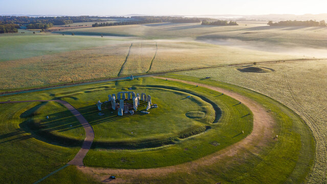 Aerial view of the ancient stone circle of Stonehenge surrounded by lush green fields and morning mist in the English countryside near Salisbury, England, United Kingdom.