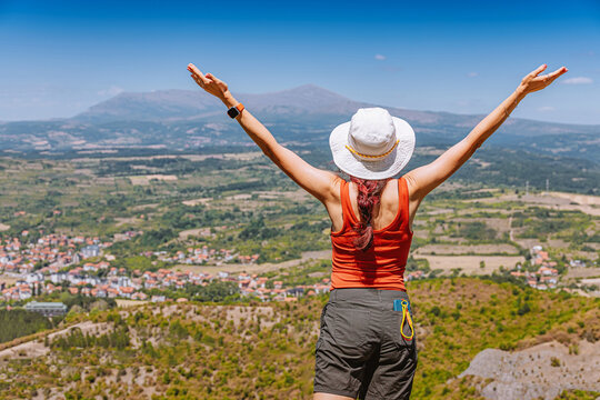 Arms raised wide, a female hiker celebrates a sunny Rtanj mountain summit above a green valley and hillside town Sokobanja. Adventure travel concept - Serbia