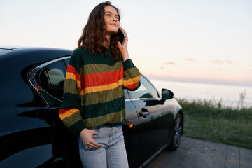 Obraz premium Smiling young woman talking on smartphone near modern car outdoors. Concept of travel, communication, and lifestyle. Soft evening lighting creates happy mood.