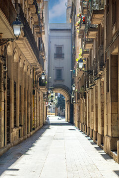 A narrow, paved street lined with historic buildings featuring wrought-iron balconies and old streetlamps, set within the picturesque atmosphere of Barcelona's Gothic Quarter.