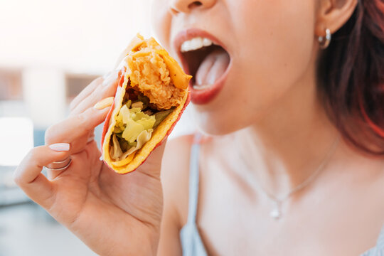 Crispy fried chicken taco with colorful shell held up to open mouth, close-up. Fast food craving concept.