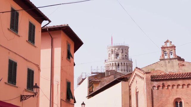 Slow gimbal walk past old town rooftops and brick church gable with Leaning Tower top peeking above in daylight, Pisa, Tuscany, Italy