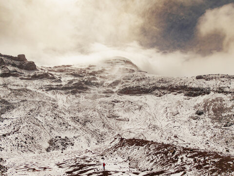 Aerial view of the snow-covered slopes of Volcano Chimborazo with a lone person in a red jacket standing in the foreground under a cloudy sky, Guano, Chimborazo, Ecuador.