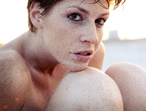 Close-up side view of a woman with short red hair and freckles sitting on a beach hugging her knees