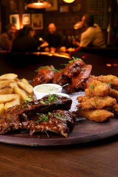 Close-up of a finger food platter featuring BBQ ribs, fried chicken, french fries, and buffalo wings on a table, with people in the background during happy hour (out of focus).
