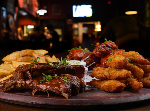 Close-up of a finger food platter featuring BBQ ribs, fried chicken, french fries, sausage, and buffalo wings, set on a table with a blurred background.