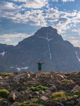 Aerial view of a hiker standing on a rocky ridge with arms raised towards the Mount of the Holy Cross, surrounded by alpine terrain in Vail, Colorado, United States.