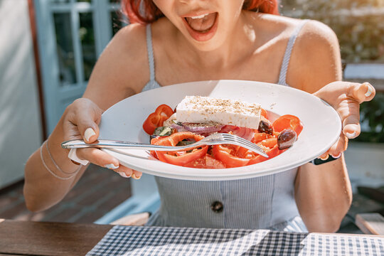Excited woman holding plate of Greek salad with feta and tomatoes at outdoor cafe table.