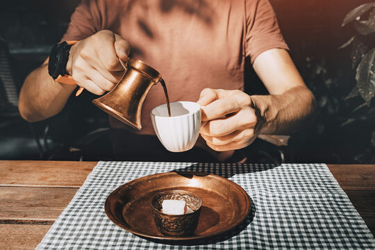 Man pouring Turkish coffee from copper cezve into white cup at wooden cafe table with checkered cloth and copper tray.