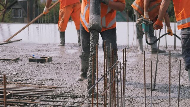 At outdoor construction site, workers wearing bright orange safety vests pour wet concrete onto steel rebar foundation. Formwork surrounds base while scaffolding and gravel spread nearby.