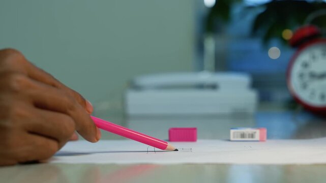 Student writing with pink pencil beside an alarm clock, Close-up of a person writing on paper with a pink pencil beside eraser and blurred alarm clock, showing study, homework, planning, or an exam.