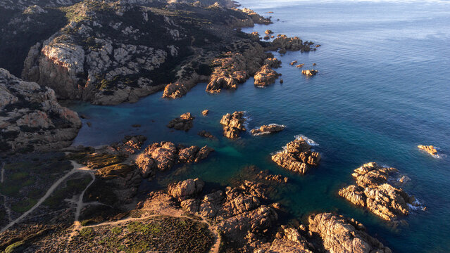 Aerial view of Cala Sarraina featuring rugged rock formations, turquoise waters, and coastal trails during golden hour in Trinita d'Agultu e Vignola, Sardinia, Italy.