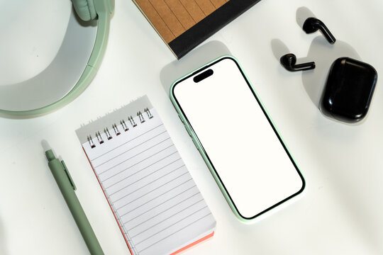 Smartphone and black wireless earbuds on white desk with notepad, pen