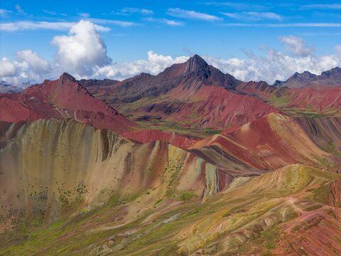 Aerial view of the colorful mineral-rich slopes of Rainbow Mountain, also known as Vinicunca, set against rugged Andean peaks under a bright blue sky with clouds in Cusco, Cuzco, Peru.