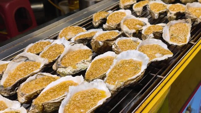 Grilled oysters with garlic topping sizzling across metal grate at outdoor street stall, showcasing Chinese street food on Hainan Island with rows of open shells cooking over charcoal