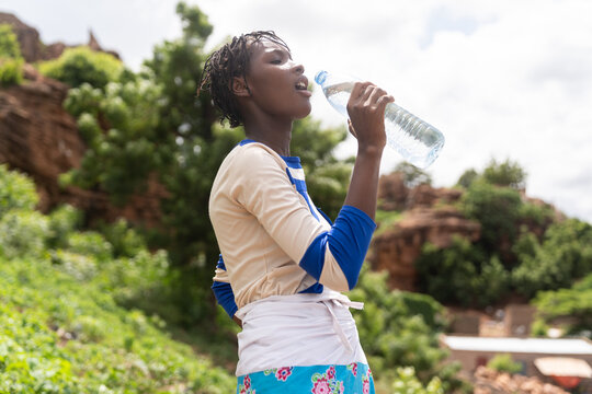 Young African woman drinks from a large water bottle outdoors against lush greenery, a natural portrait about hydration, health and access to clean water